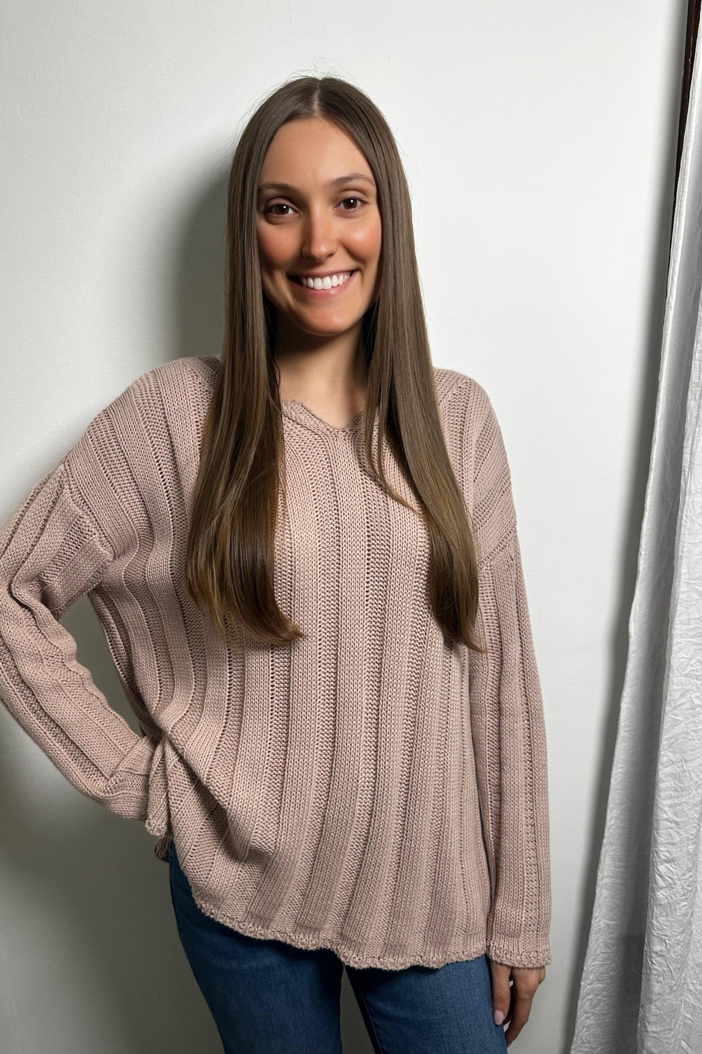 Woman wearing a beige sweater and blue jeans against a white wall.