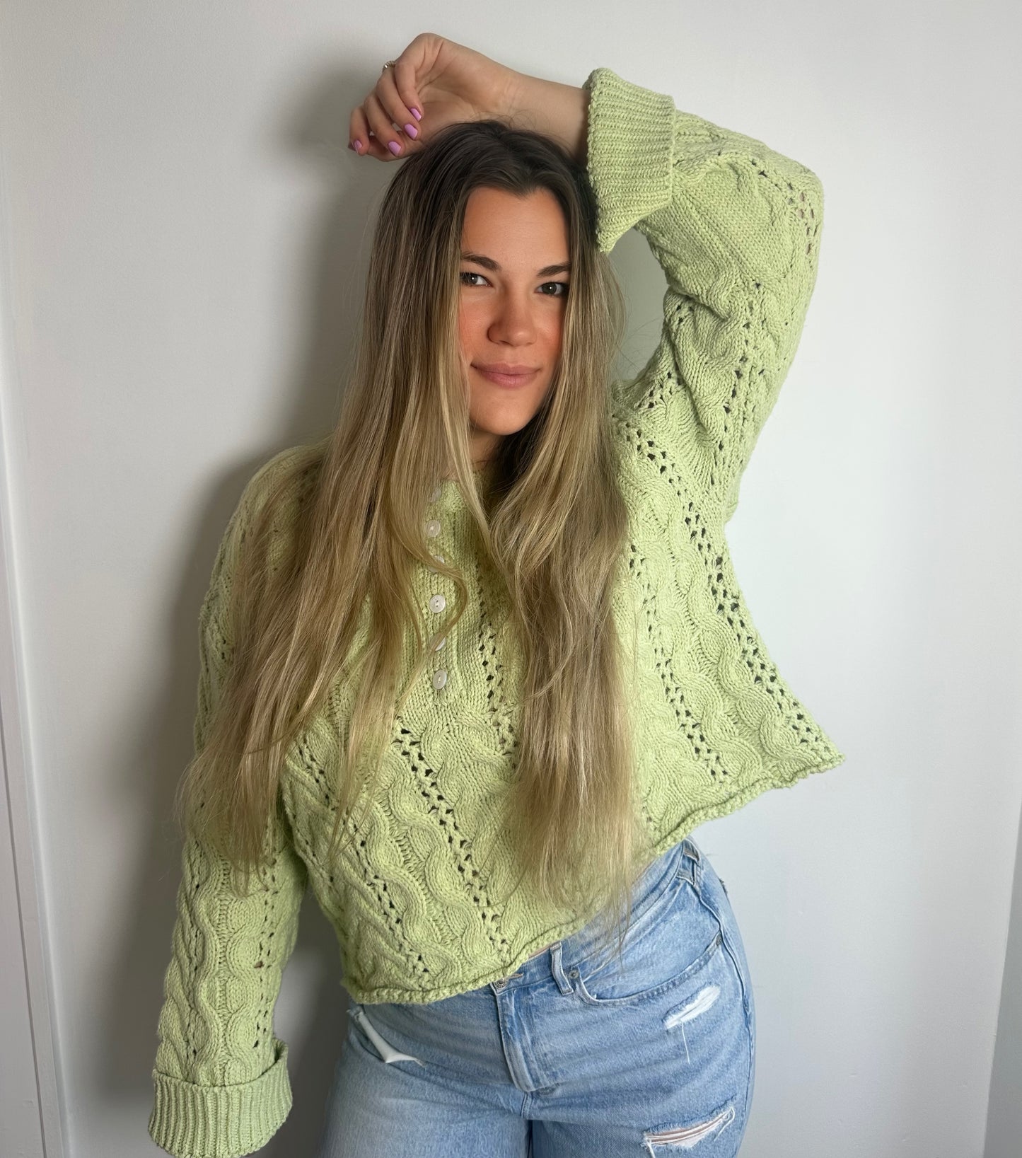 Woman wearing a light green knitted sweater and blue jeans against a white wall.
