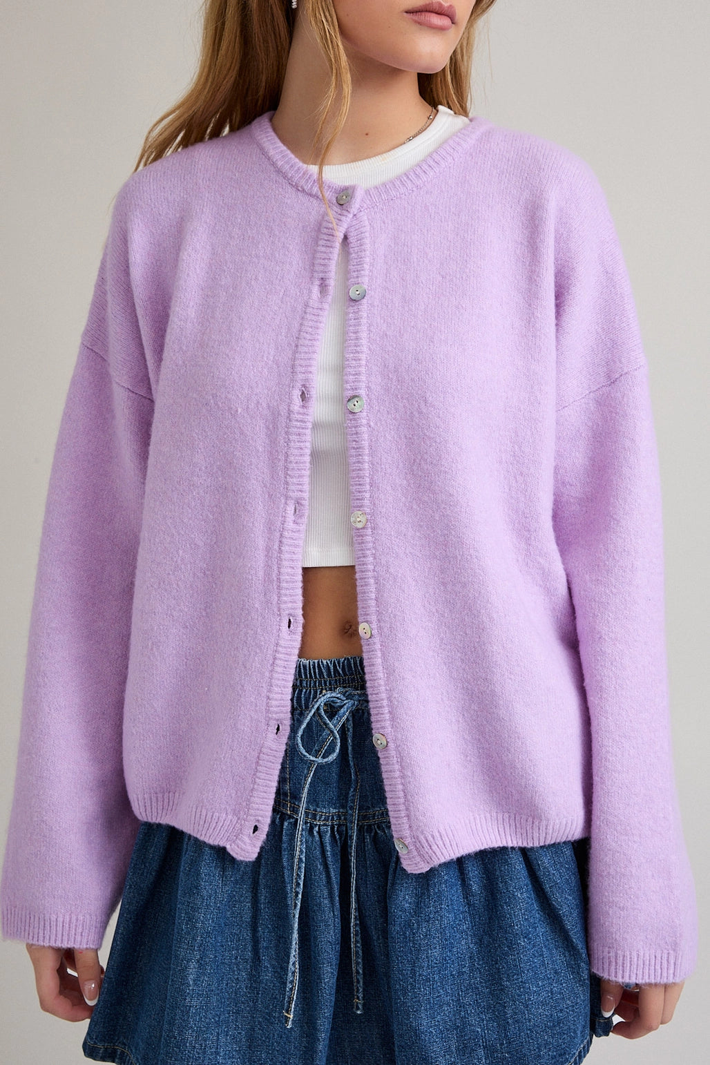 Woman wearing a light purple cardigan over a white top and blue denim skirt on a plain background
