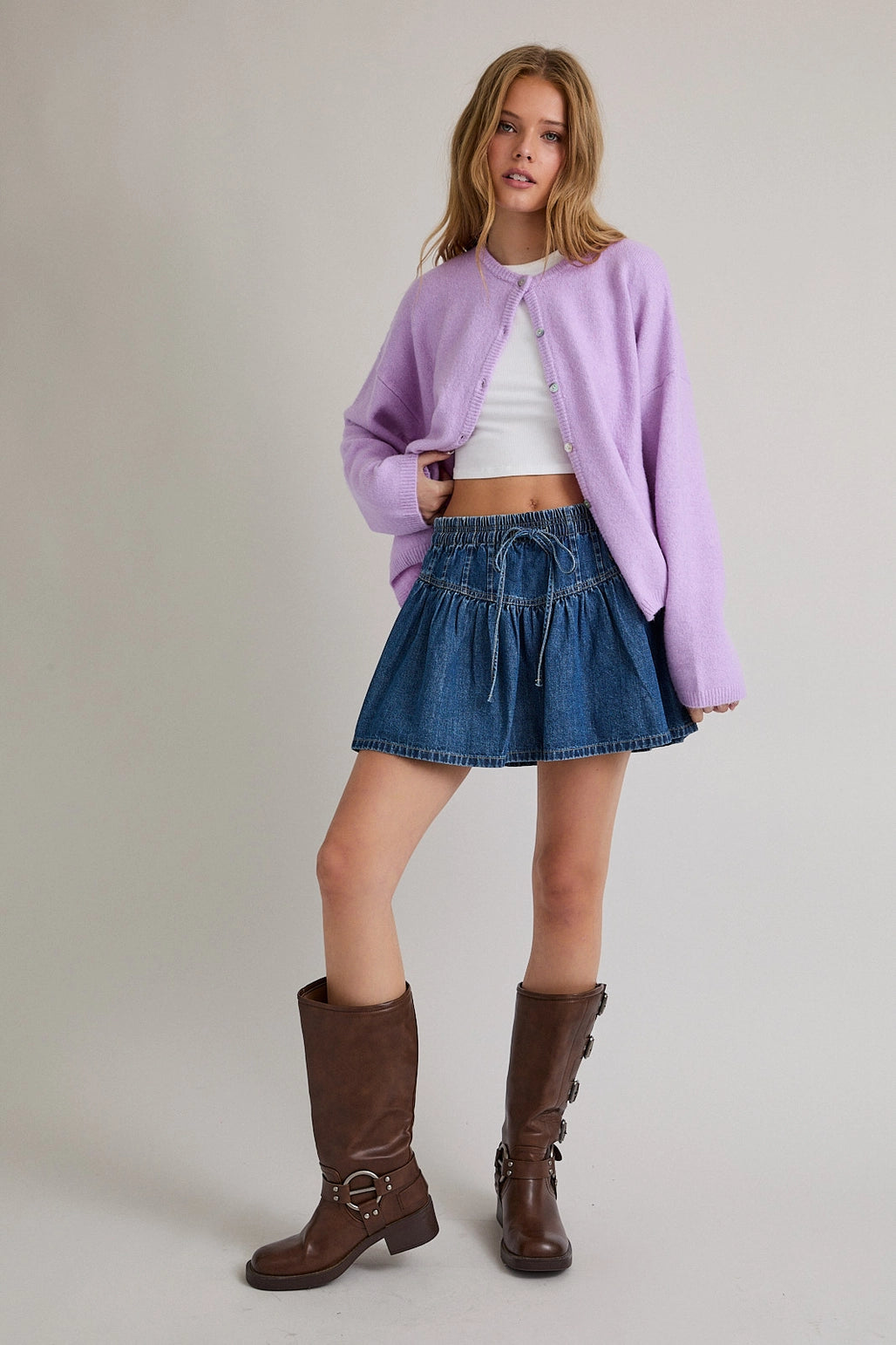 Woman wearing a light purple cardigan over a white top and blue denim skirt on a plain background