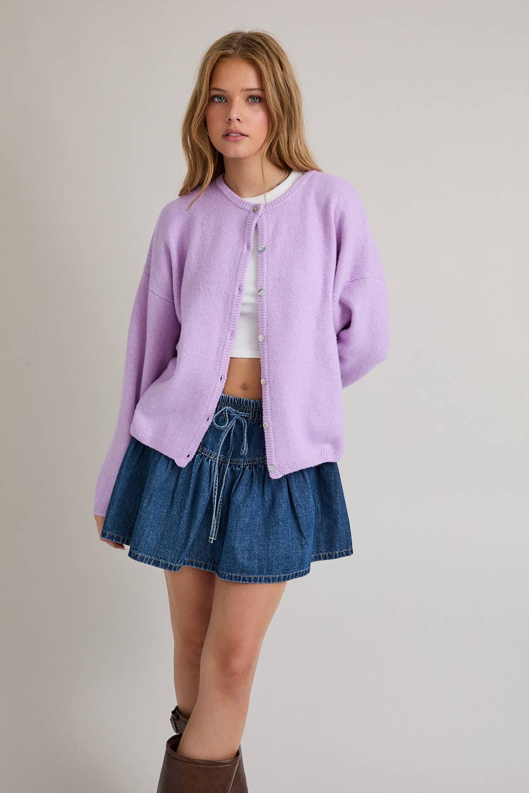Woman wearing a light purple cardigan over a white top and blue denim skirt on a plain background