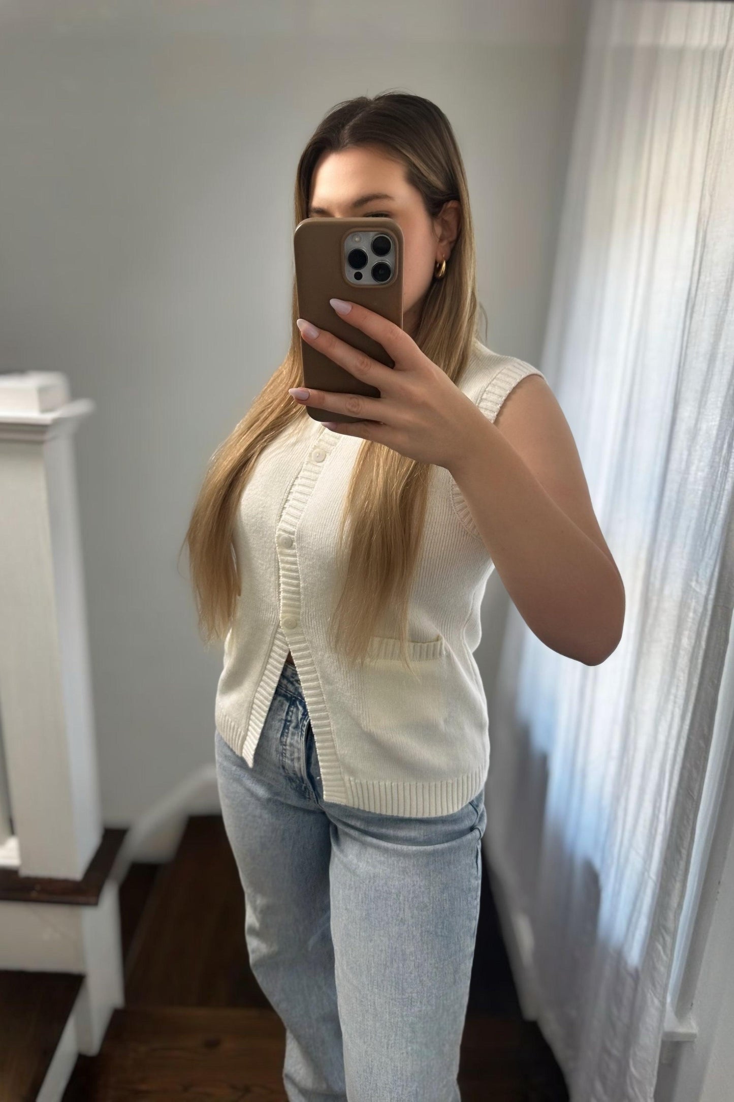 Person wearing a white sleeveless sweater and light blue jeans taking a mirror selfie.