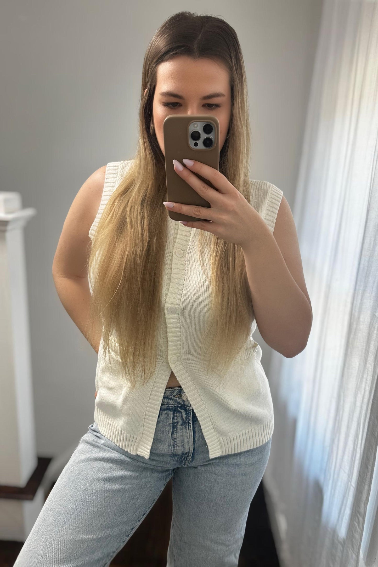Woman taking a mirror selfie wearing a white sleeveless cardigan and blue jeans.