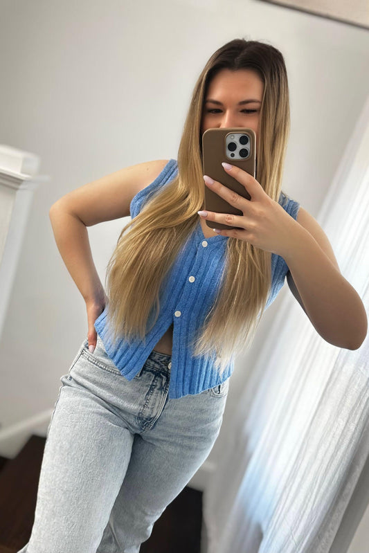 Woman taking a mirror selfie wearing a blue sleeveless cardigan and light jeans.