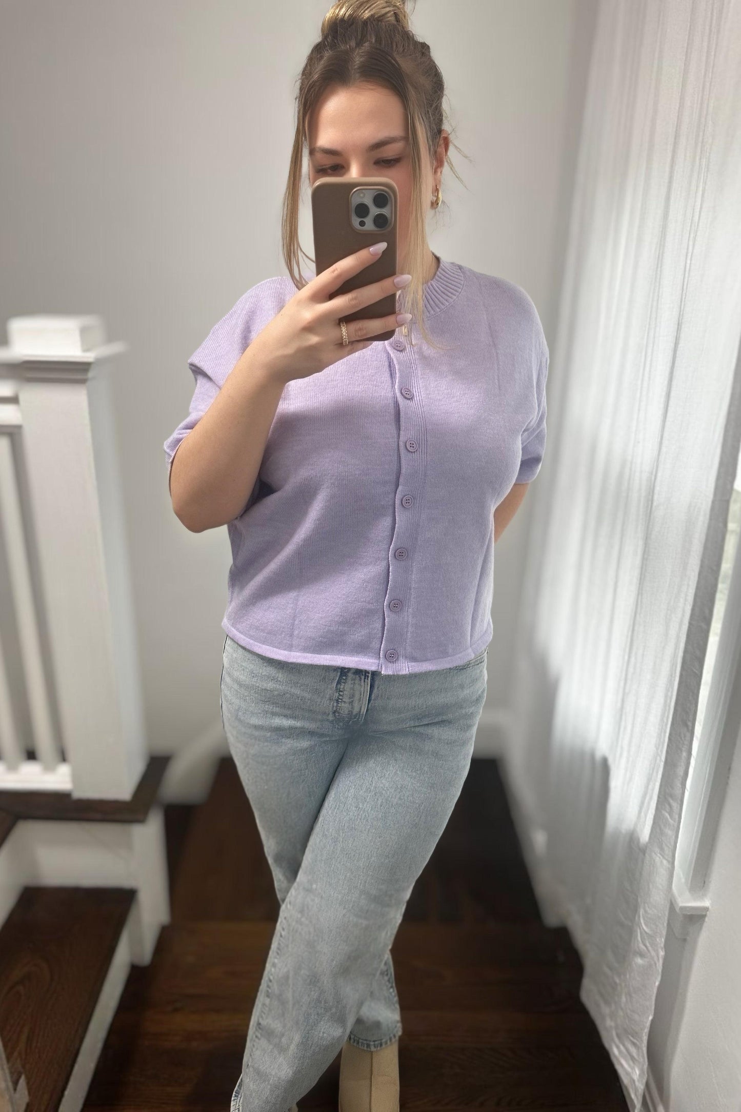 Woman taking a mirror selfie wearing a light purple shirt and light blue jeans in a room with white walls and wooden floor.