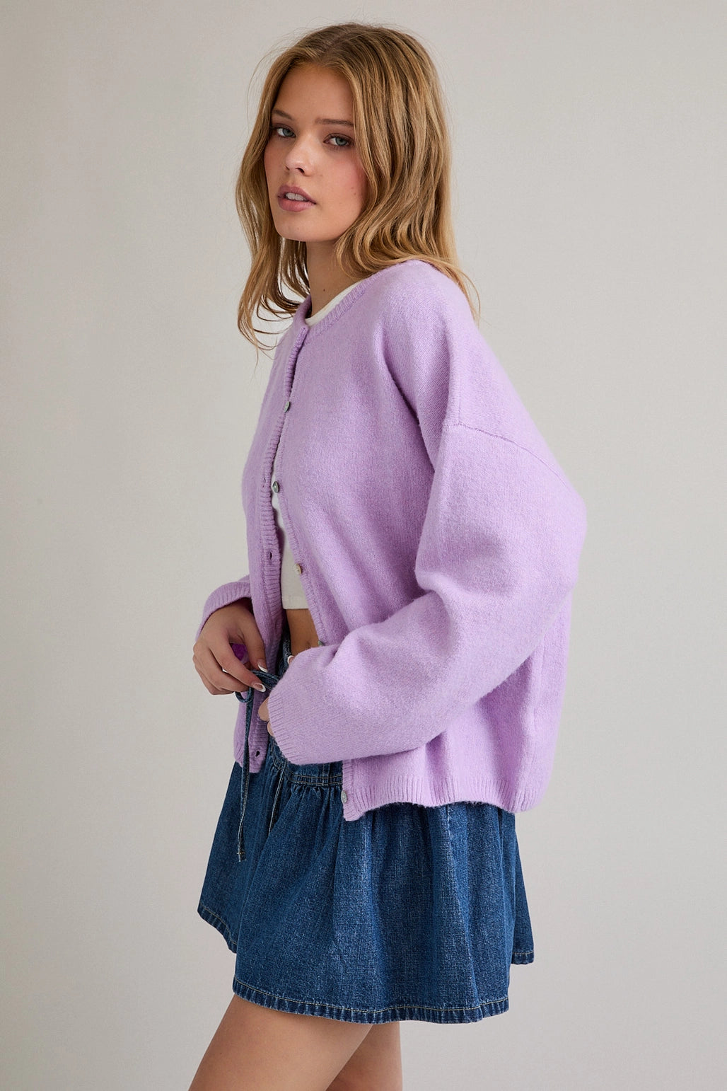 Woman wearing a light purple cardigan over a white top and blue denim skirt on a plain background