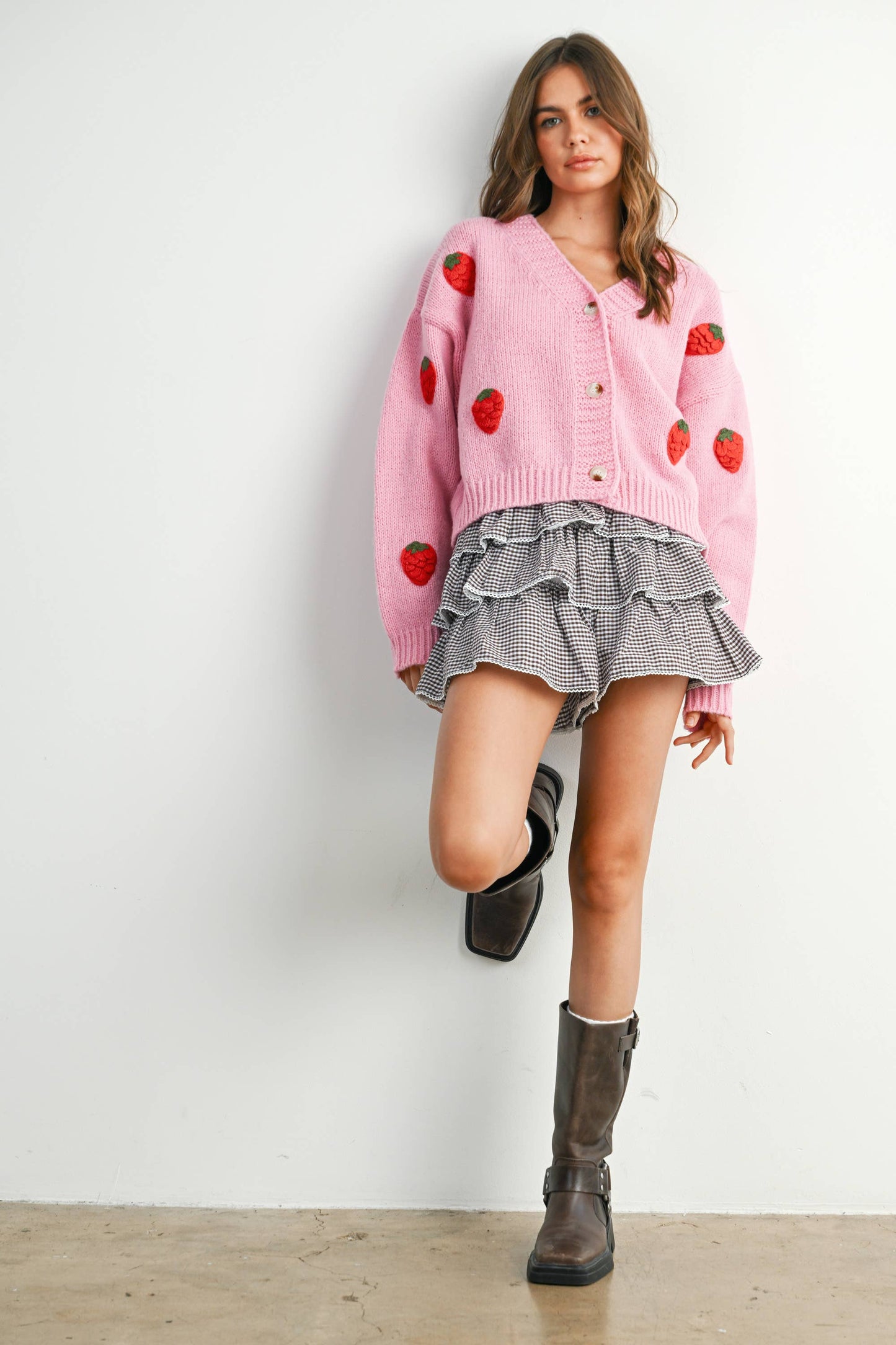 Woman wearing a pink cardigan with red strawberry patterns, a gray ruffled skirt, and brown boots against a white wall.